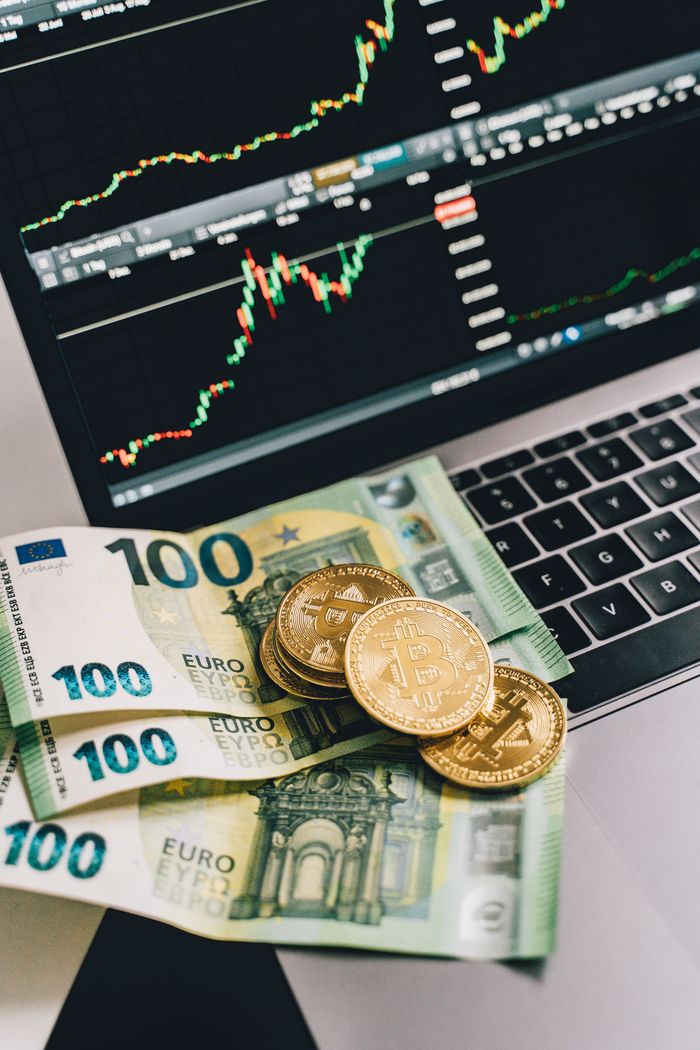 Bitcoin coins and Euro notes on a laptop with a stock market graph display, symbolizing modern finance.